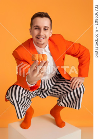 extravagant funny young man in orange jacket and striped pants holding cupcake on orange background. extravagant funny young man in orange jacket and striped pants holding cupcake on orange background. 109636732
