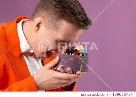 birthday man in orange jacket bites cake on purple background birthday man in orange jacket bites cake on purple background 109636754