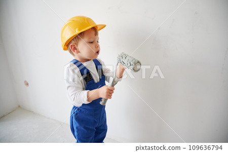 Cute child construction worker standing by the wall and holding paint roller. Kid in safety helmet and work overalls using painting tool while working on home renovation. Cute child construction worker standing by the wall and holding paint roller. Kid in safety helmet and work overalls using painting tool while working on home renovation. 109636794