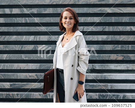 Cheerful middle-aged woman in stylish clothes looking at the camera while standing outdoors. Female in coat with leather folder. Woman with ginger hair wearing stylish clothes. Cheerful middle-aged woman in stylish clothes looking at the camera while standing outdoors. Female in coat with leather folder. Woman with ginger hair wearing stylish clothes. 109637525