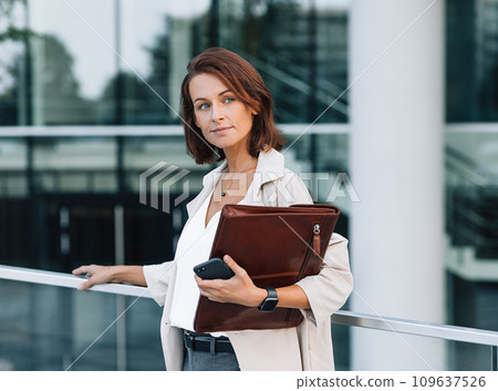 Confident middle-aged woman in stylish clothes standing outdoors. Female with ginger hair holding a leather folder while standing against an office building. Confident middle-aged woman in stylish clothes standing outdoors. Female with ginger hair holding a leather folder while standing against an office building. 109637526