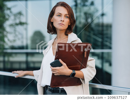 Confident middle-aged businesswoman looking away. Female in formal clothes holding leather folder while standing outdoors. Confident middle-aged businesswoman looking away. Female in formal clothes holding leather folder while standing outdoors. 109637527