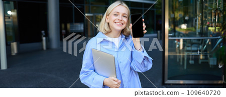 Happy smiling woman showing smartphone screen, demonstrating mobile phone display, standing in college outdoors, posing near campus with laptop and telephone 109640720