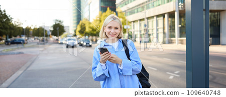 Street style shot of young smiling woman with smartphone, standing on street, bright sunny day, road with cars behind her back, using mobile phone, waiting for someone outdoors 109640748
