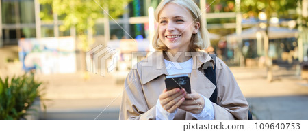 Carefree smiling woman standing on sunny street, using mobile phone, waiting for car or bus with smartphone, looking around Carefree smiling woman standing on sunny street, using mobile phone, waiting for car or bus with smartphone, looking around 109640753