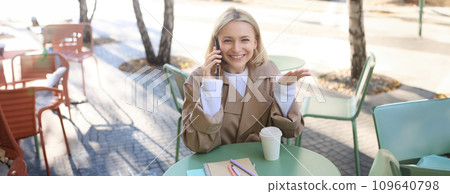 Happy blond female model, sitting outdoors in cafe, answer phone call, having conversation on smartphone, laughing and smiling Happy blond female model, sitting outdoors in cafe, answer phone call, having conversation on smartphone, laughing and smiling 109640798