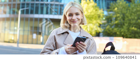 Close up portrait of happy blond girl waiting outside, standing on street with smartphone, looking at camera and smiling 109640841
