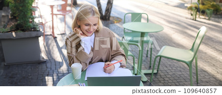 Image of smiling young female student, working on project, sitting outside on sunny day in cafe, looking at laptop and making notes, studying, doing homework outdoors Image of smiling young female student, working on project, sitting outside on sunny day in cafe, looking at laptop and making notes, studying, doing homework outdoors 109640857
