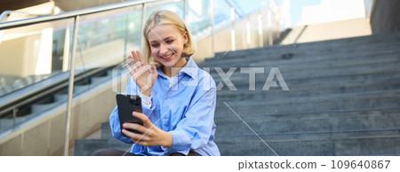 Cute smiling young woman, sitting on stairs with mobile phone, waving hand and saying hello during video chat, connects to online meeting 109640867