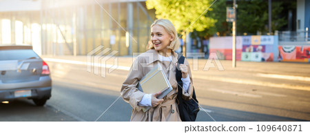 Lifestyle portrait of young woman walking on street with backpack and notebook, looking aside, saying hello to someone on her way to university or college Lifestyle portrait of young woman walking on street with backpack and notebook, looking aside, saying hello to someone on her way to university or college 109640871