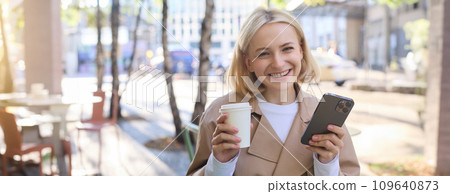 Portrait of blond smiling woman with smartphone, holding cup of coffee, drinking chai and enjoying sunny day outdoors in city centre 109640873