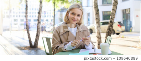 Portrait of carefree, blond cute woman working with documents, drinking takeaway coffee and sitting in cafe outdoors with documents, writing essay, student doing homework Portrait of carefree, blond cute woman working with documents, drinking takeaway coffee and sitting in cafe outdoors with documents, writing essay, student doing homework 109640889