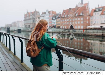 Attractive young female tourist is exploring new city. Redhead girl holding a mobile phone on city street in Gdansk. Traveling Europe in autumn. Vacation concept, rear view Attractive young female tourist is exploring new city. Redhead girl holding a mobile phone on city street in Gdansk. Traveling Europe in autumn. Vacation concept, rear view 109641053