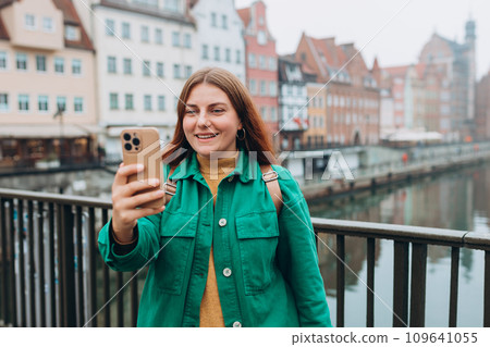 Happy 30s Women taking selfie on urban background. Young beautiful girl say Hi. Gdansk old town and famous Zuraw crane, A beautiful redhead woman is standing near Motlawa river, Traveler 109641055