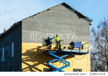 Residential house facade painting works after siding with wooden planks using scissor lift platform Residential house facade painting works after siding with wooden planks using scissor lift platform 109641428