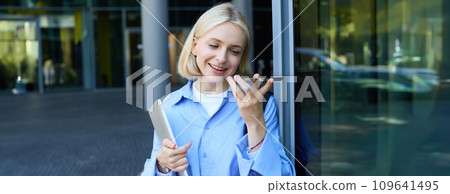 Portrait of beautiful, smiling young student, woman records voice message, talking on speakerphone, standing near office building on street and holding laptop with backpack Portrait of beautiful, smiling young student, woman records voice message, talking on speakerphone, standing near office building on street and holding laptop with backpack 109641495