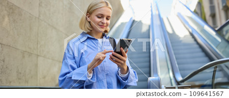 Portrait of young stylish woman, female employee, standing near escalator, using mobile phone, sending message on smartphone, tap screen, smiling and reading on telephone 109641567