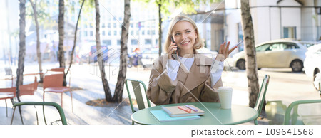 Image of stylish blond woman talking on mobile phone, drinking coffee in cafe outdoors, enjoying warm weather in city centre, answer telephone call on smartphone Image of stylish blond woman talking on mobile phone, drinking coffee in cafe outdoors, enjoying warm weather in city centre, answer telephone call on smartphone 109641568