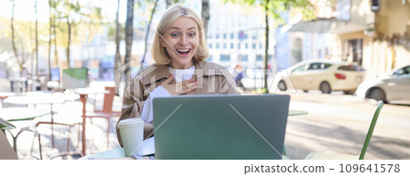 Lifestyle portrait of young surprised woman, looking at laptop with amazed face, sitting in coffee shop with excited face expression, spending time outdoors on sunny day Lifestyle portrait of young surprised woman, looking at laptop with amazed face, sitting in coffee shop with excited face expression, spending time outdoors on sunny day 109641578
