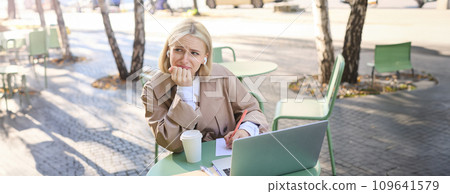 Lifestyle and technology concept. Young blond working woman in trench coat, studying, working on project on laptop, sitting in coffee shop on sunny day, making notes, attending online course 109641579