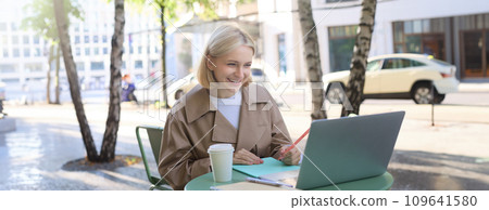 Young woman, freelancer, student doing homework in outdoor cafe, drinking her coffee on street, using laptop, connects to online lecture or doing course in internet 109641580
