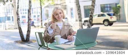 Remote workplace. Smiling young woman freelancer, student using laptop in outdoor cafe, talking to someone, attends online lecture, drinking coffee Remote workplace. Smiling young woman freelancer, student using laptop in outdoor cafe, talking to someone, attends online lecture, drinking coffee 109641604