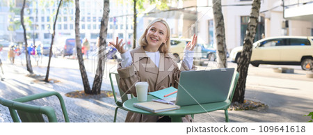 Image of young modern woman, chatting with someone online, having video call via laptop, sitting in outdoor cafe, drinking coffee, gesturing and smiling Image of young modern woman, chatting with someone online, having video call via laptop, sitting in outdoor cafe, drinking coffee, gesturing and smiling 109641618
