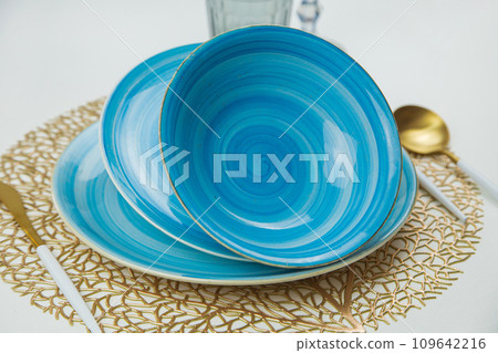 Bright blue serving plates with a spiral pattern, tilted one after the other, serving devices of gold - white color are located on the side, liquid glasses are visible behind them, a close-up side Bright blue serving plates with a spiral pattern, tilted one after the other, serving devices of gold - white color are located on the side, liquid glasses are visible behind them, a close-up side 109642216