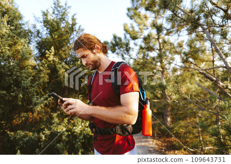 Hiker man with backpack checks the route on his phone on the walking trail Hiker man with backpack checks the route on his phone on the walking trail 109643731