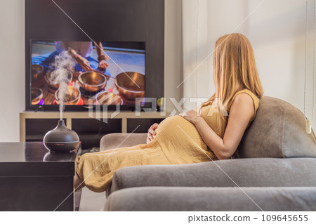 A blissful pregnant woman immerses in relaxation, savoring the soothing aroma from a diffuser while indulging in a calming TV video, embracing tranquility during her pregnant journey A blissful pregnant woman immerses in relaxation, savoring the soothing aroma from a diffuser while indulging in a calming TV video, embracing tranquility during her pregnant journey 109645655