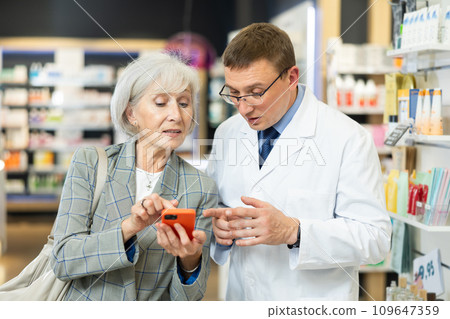 Male pharmacist carefully looks at photo in mature woman clients phone Male pharmacist carefully looks at photo in mature woman clients phone 109647359