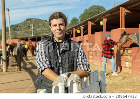 Portrait of positive adult man landowner wearing plaid shirt and posing during daytime on yard of horse stables 109647418
