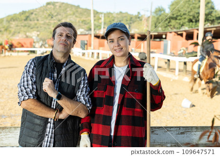 Experienced man and woman in plaid shirts looking at camera while holding working tool on horse ranch in spring Experienced man and woman in plaid shirts looking at camera while holding working tool on horse ranch in spring 109647571