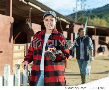 Portrait of young woman with brushes in her hands for cleaning and caring for horses at horse farm Portrait of young woman with brushes in her hands for cleaning and caring for horses at horse farm 109647889