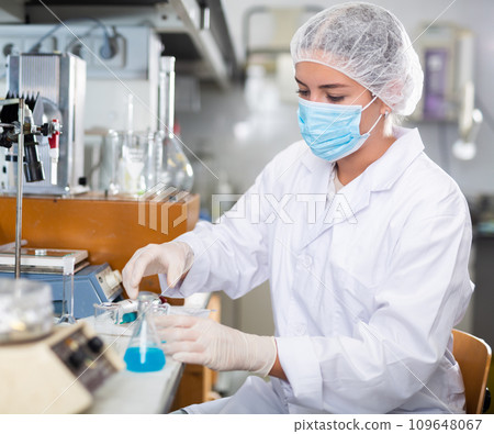 Skillful young female lab assistant in white coat pouring blue liquid reagent into beaker to create chemical solution Skillful young female lab assistant in white coat pouring blue liquid reagent into beaker to create chemical solution 109648067