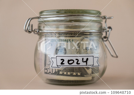 Saving Money In Glass Jar filled with Dollars banknotes. 2024 year transcription in front of jar. Managing personal finances extra income for future insecurity 109648710