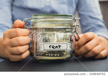 Female hands holding Glass jar full of American currency dollars cash banknote with text RENT. Preparation saving money. Moderate consumption and economy Female hands holding Glass jar full of American currency dollars cash banknote with text RENT. Preparation saving money. Moderate consumption and economy 109648742