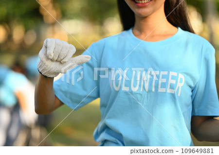 Cropped shot of female pointing to volunteer uniform and smiling over nature background Cropped shot of female pointing to volunteer uniform and smiling over nature background 109649881