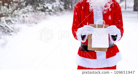Santa Claus outdoor in winter and snow handing in hand eco paper bags with craft gift, food delivery. Shopping, packaging recycling, handmade, delivery for Christmas and New year 109651947