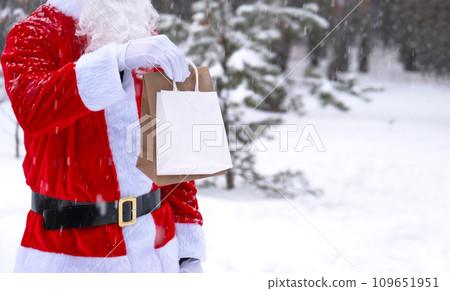 Santa Claus outdoor in winter and snow handing in hand eco paper bags with craft gift, food delivery. Shopping, packaging recycling, handmade, delivery for Christmas and New year 109651951