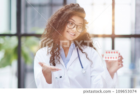 Young pharmacist woman holding pills while sitting at the table in the office Young pharmacist woman holding pills while sitting at the table in the office 109653074
