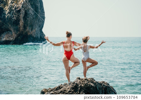 Woman and her daughter practicing balancing yoga pose on one leg up together on rock in the sea. Silhouette mother and daughter doing yoga at beach 109653784