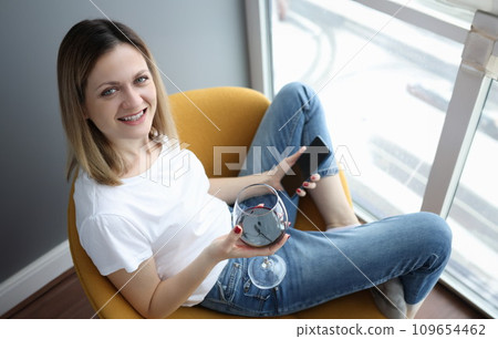 Young woman sitting on armchair with glass of red wine and phone 109654462