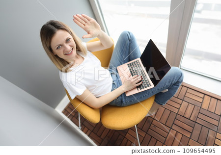 Young woman sitting on armchair with laptop and waving hand Young woman sitting on armchair with laptop and waving hand 109654495