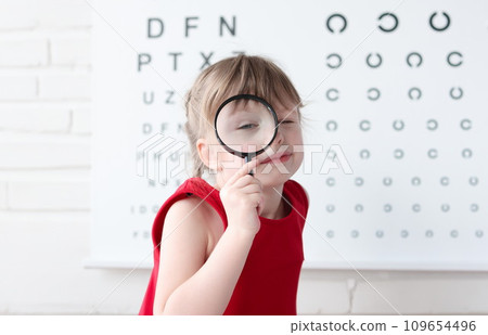 Little girl with magnifying glass standing on background of table for eye test 109654496