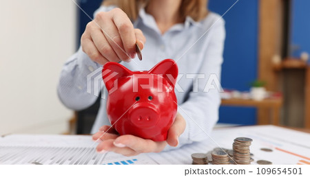 Business woman throwing coins into piggy bank at table with graphs closeup Business woman throwing coins into piggy bank at table with graphs closeup 109654501