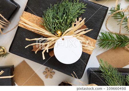 Black Christmas present with blank gift tag, with fir branch and decorations, Mockup 109660498