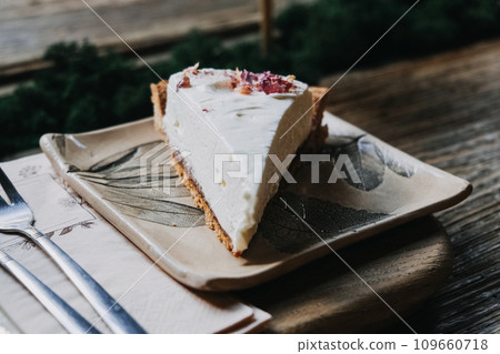 Creamy Cheesecake Slice on Botanical Ceramic Plate. Elegant cheesecake slice topped with dried petals, perfectly presented on a leaf-patterned ceramic plate. 109660718