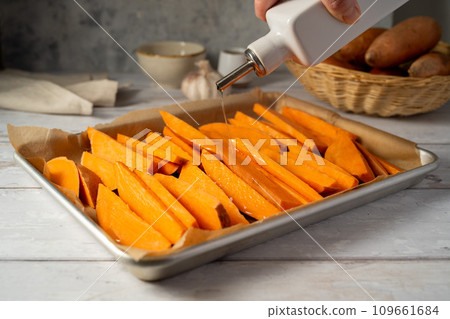 Cooking sweet potato wedges, fries on a baking pan. Hand adding oil. Cooking sweet potato wedges, fries on a baking pan. Hand adding oil. 109661684