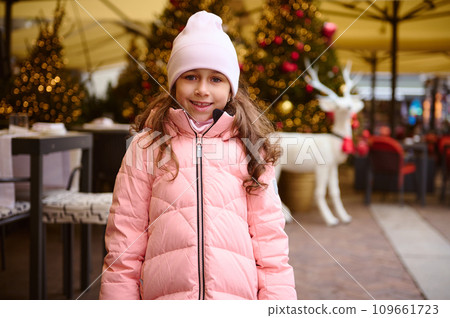 Lovely happy little kid girl smiles at camera, standing outdoors at Christmas fair, enjoying the upcoming festive events 109661723